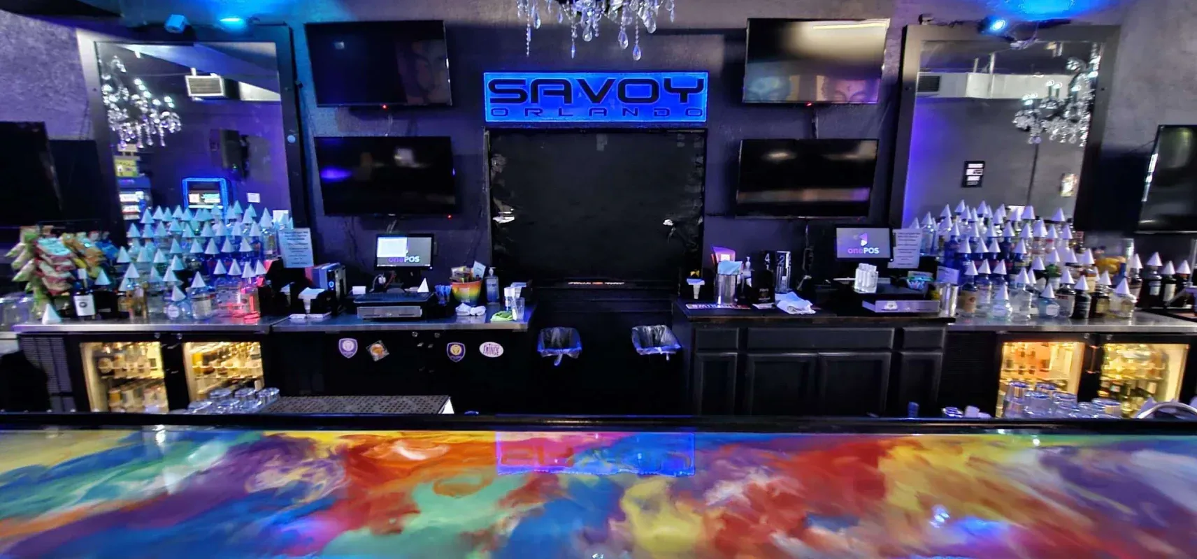 Neon-lit nightclub bar with blue signage, mirrored shelves, and colorful floor lights