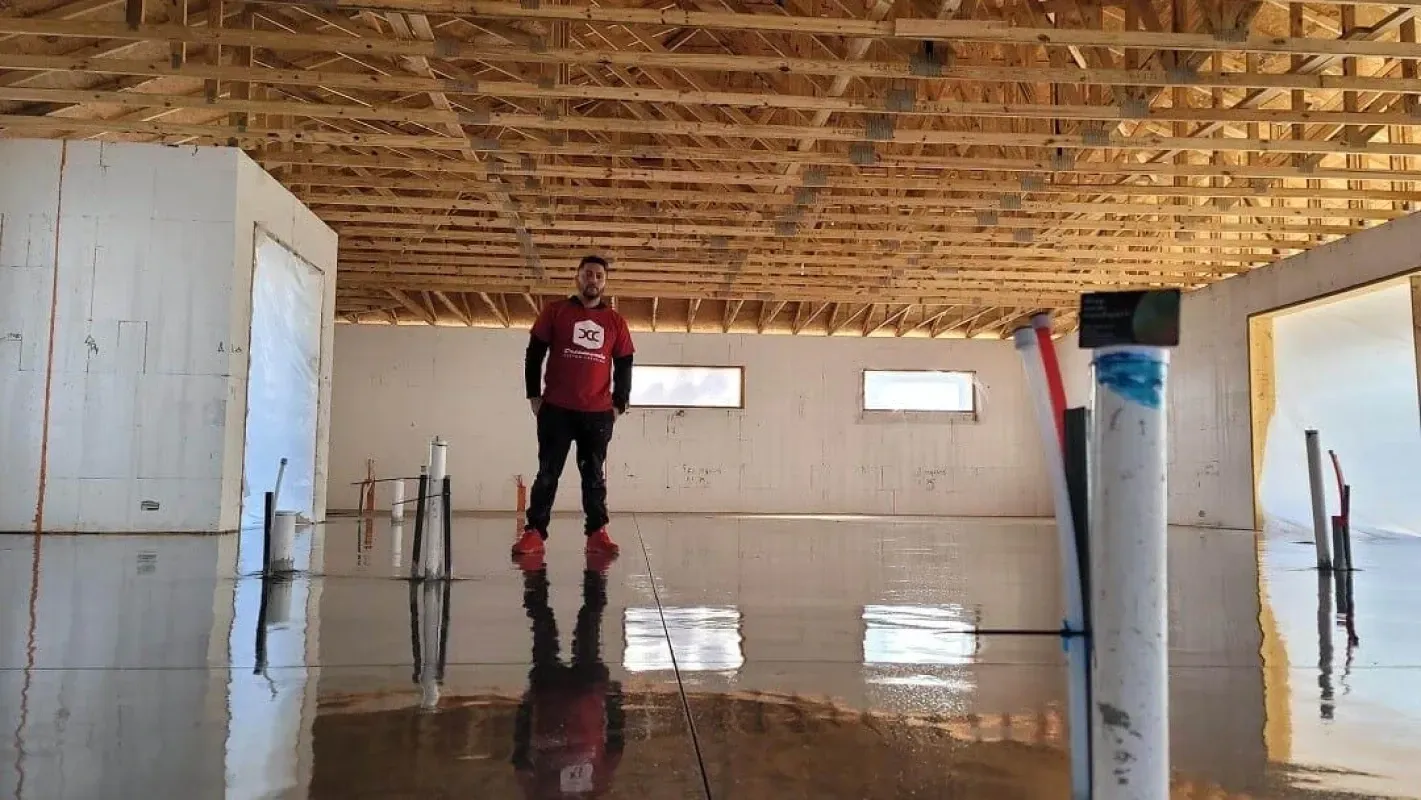Person standing in a flooded room with reflective water, unfinished wooden ceiling, and white walls