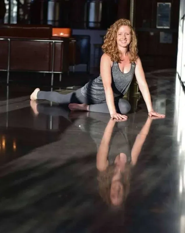Woman in a black workout outfit posing on a glossy studio floor, smiling with her reflection visible.