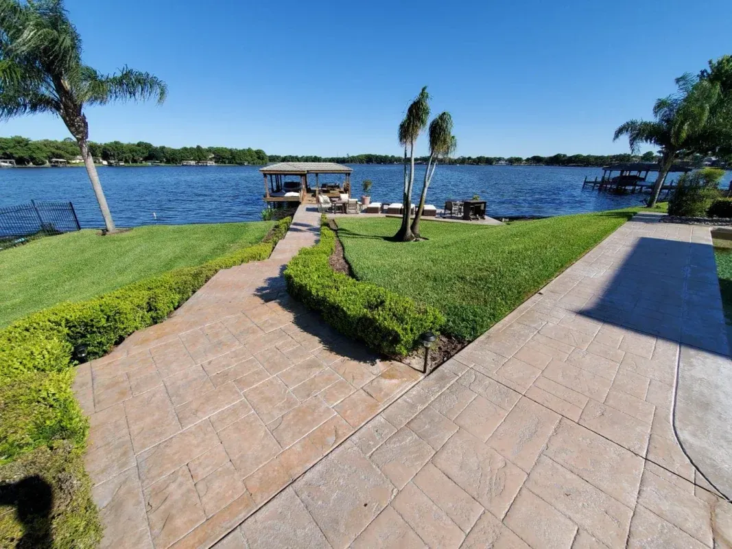 Tiled walkway to a dock on a sunny lakeside lawn with palm trees and blue water
