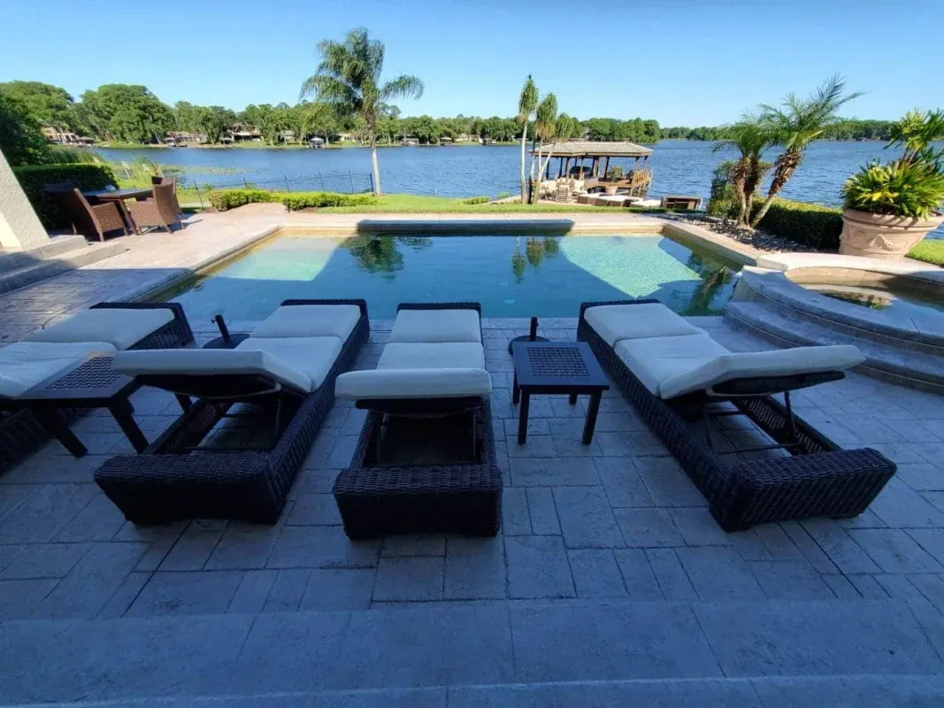 Poolside lounge chairs facing a lake with palm trees and blue sky in the background