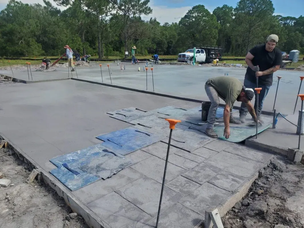 Workers smoothing fresh concrete on a slab outdoors with tools and orange markers nearby