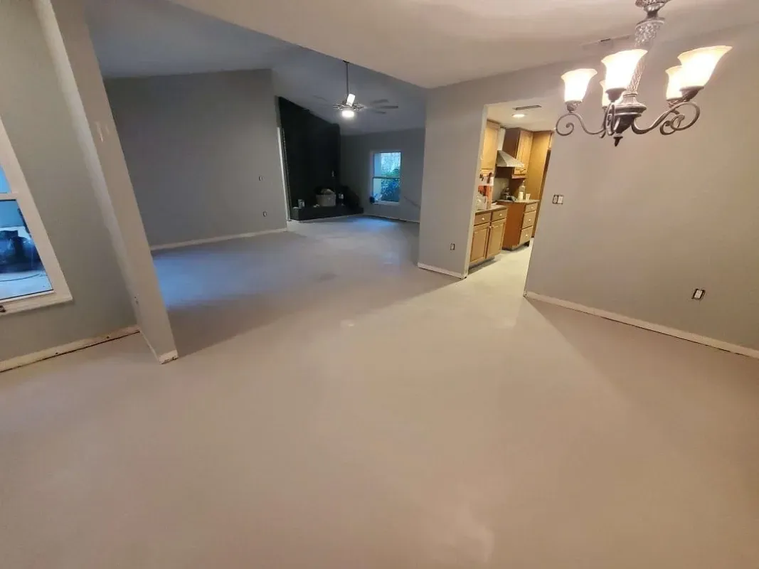Empty beige living room with carpet, open doorway, and chandelier-lit dining area beyond