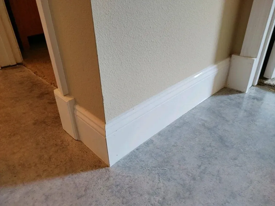 Hallway corner with beige walls, white baseboard, and carpet meeting gray flooring at a doorway