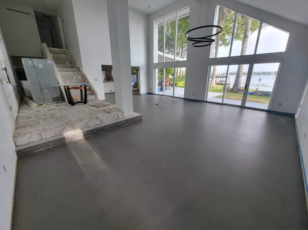 Empty modern living room with large windows, gray floor, and a staircase leading to an open kitchen area