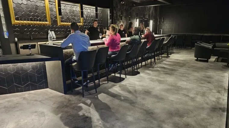 Three people sit at a bar in a dimly lit lounge with dark decor and a concrete floor.