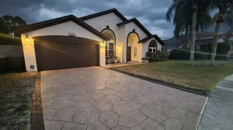 Single-story suburban house with lit entrance, dark garage door, and a wide stamped concrete driveway at dusk