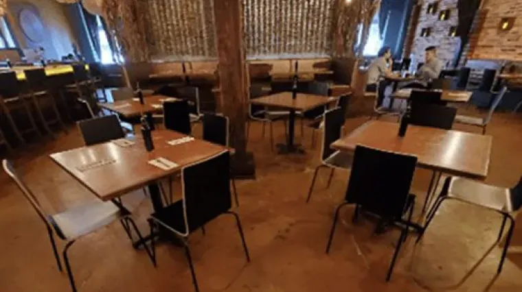 Empty restaurant dining area with wooden tables and chairs, dim warm lighting, and a bar in the background