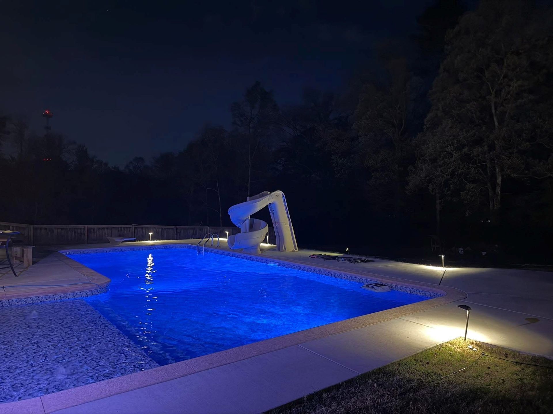 Nighttime backyard pool glowing blue with a white slide and landscape lights surrounding it