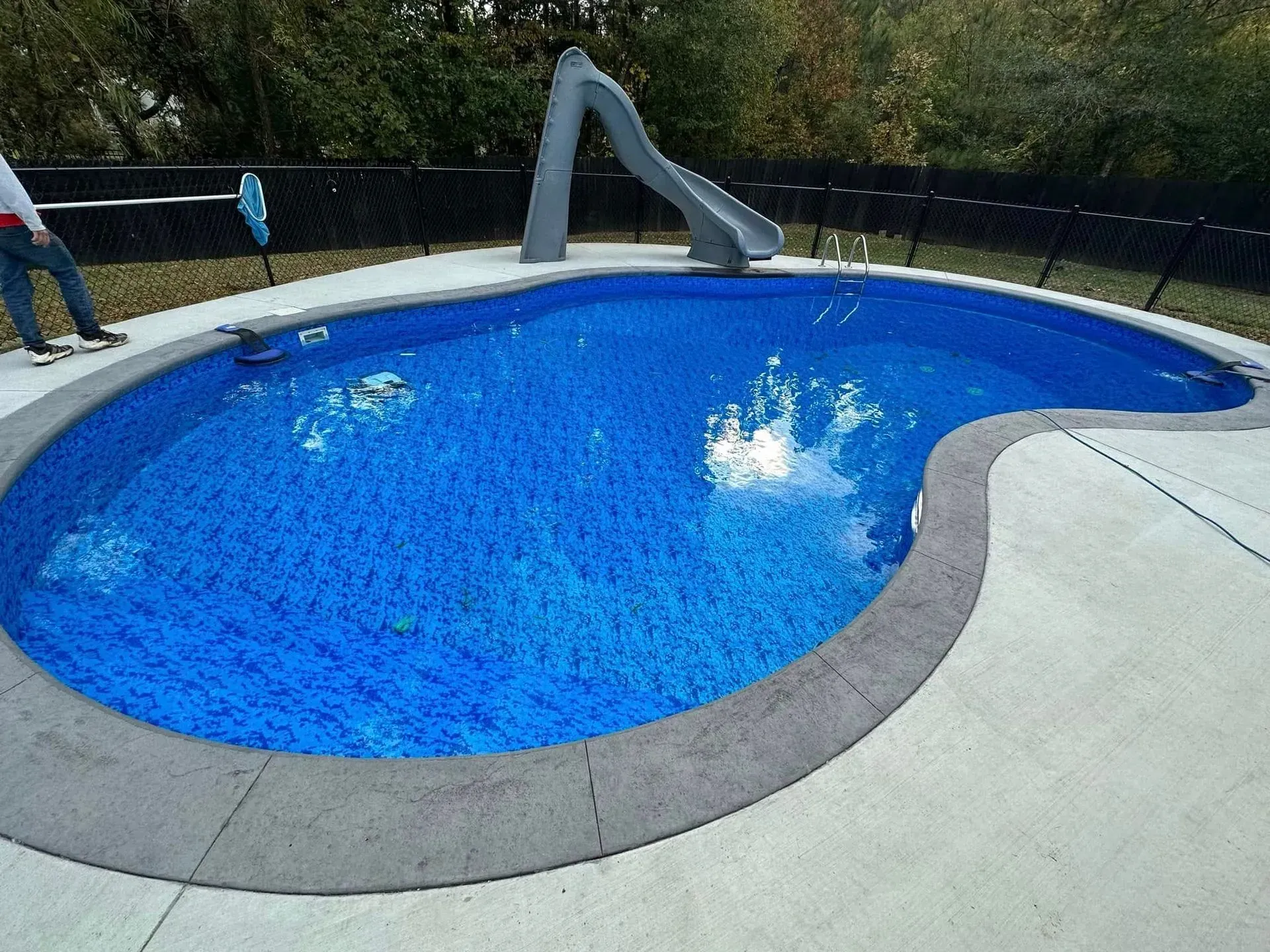 Blue-tiled pool with concrete surround; a gray slide; a black fence and a person in a yard.