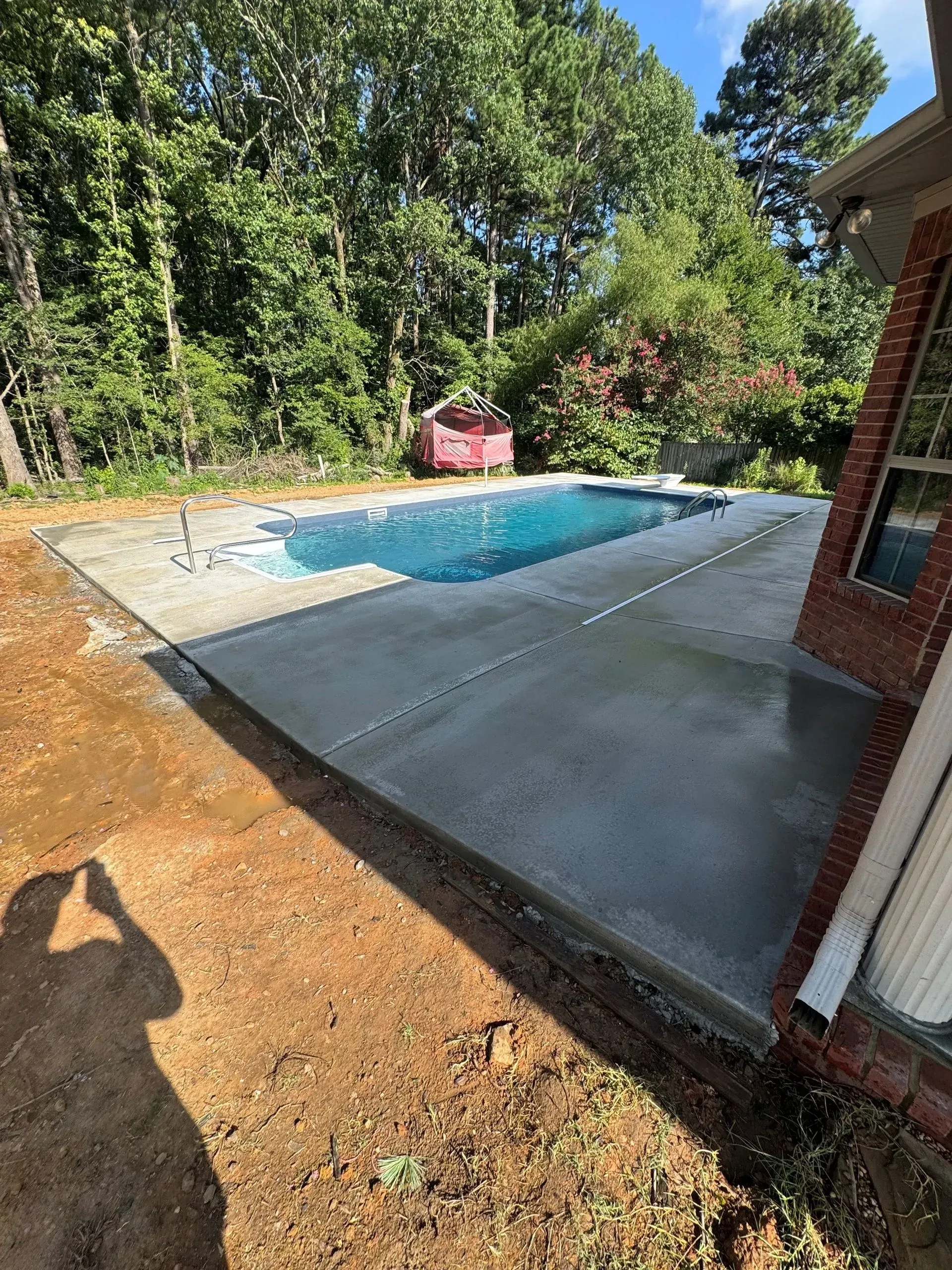 Newly constructed concrete patio surrounds a rectangular swimming pool, near a brick house and trees.