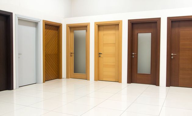 Row of six different wooden doors displayed against a white wall on a glossy tiled floor.
