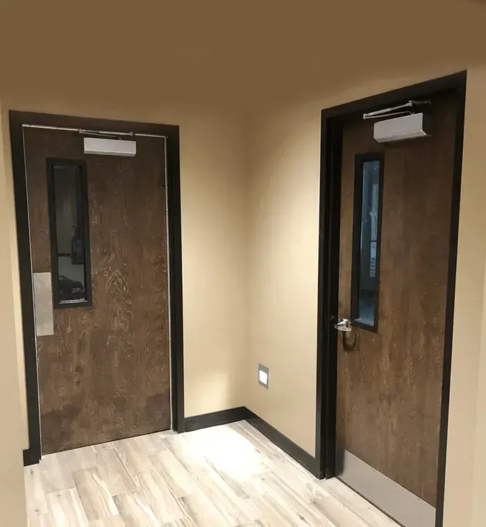Two brown doors with narrow windows in a beige hallway with light wood flooring.