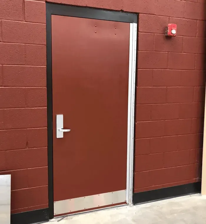 Red door in a brick wall with a silver handle and a red alarm light above it.
