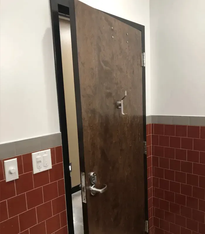 Open wooden restroom door in a tiled hallway, slightly ajar, with wall switches beside it.