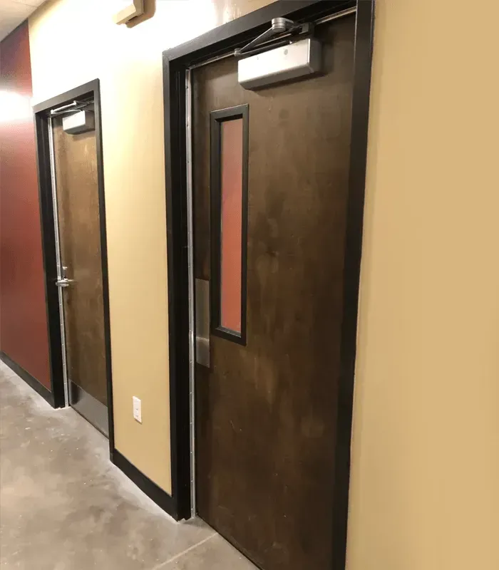 Two dark brown doors with narrow windows in a beige hallway with concrete floors
