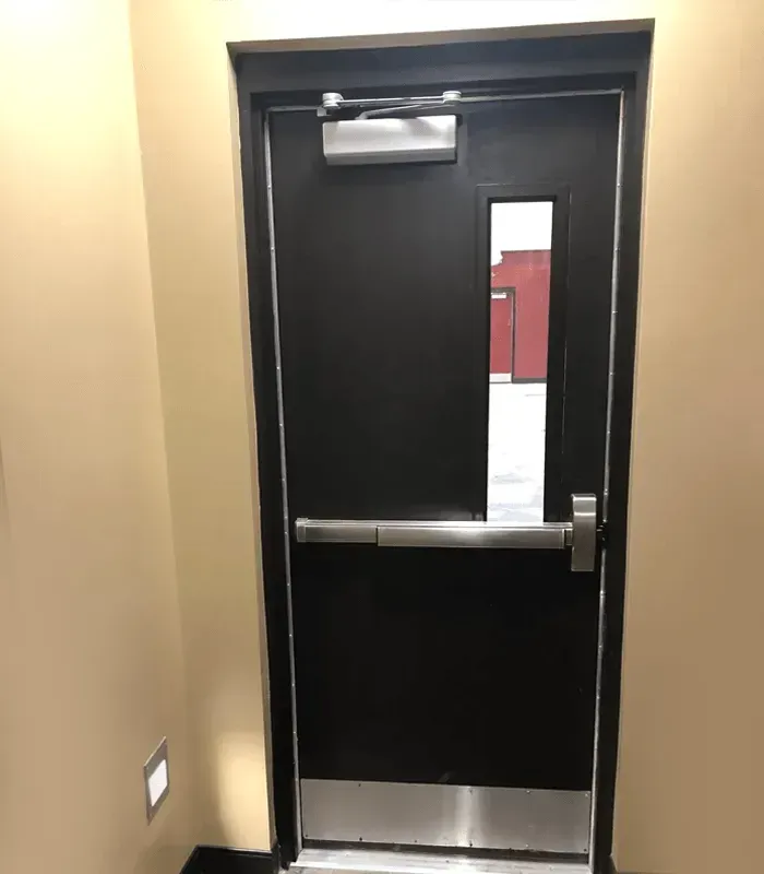 Black door with push bar and narrow window in a beige hallway