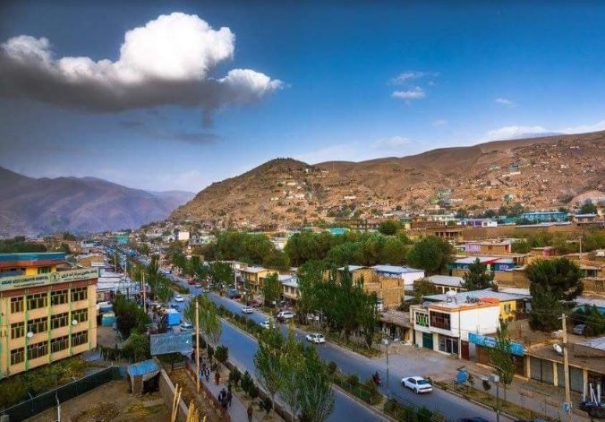 An aerial view of a town nestled in a valley with a road, buildings, and mountains under a blue sky with clouds.