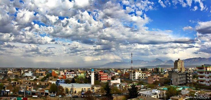 A cityscape under a cloudy blue sky. Buildings of various colors and heights spread across the landscape, with distant mountains in the background.