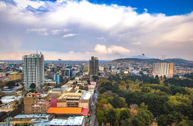 Cityscape of Kabul, Afghanistan, under a cloudy sky. Tall buildings, trees, and a distant hill with a flag are visible.