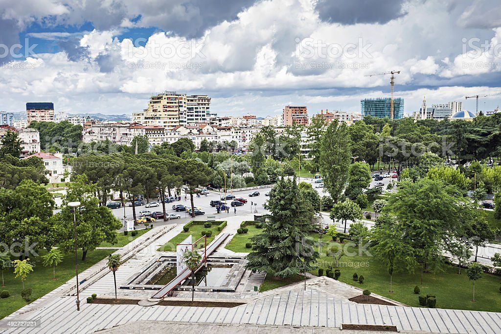 Park with trees, walkways, and a city skyline under a cloudy sky. Buildings of various heights fill the background.