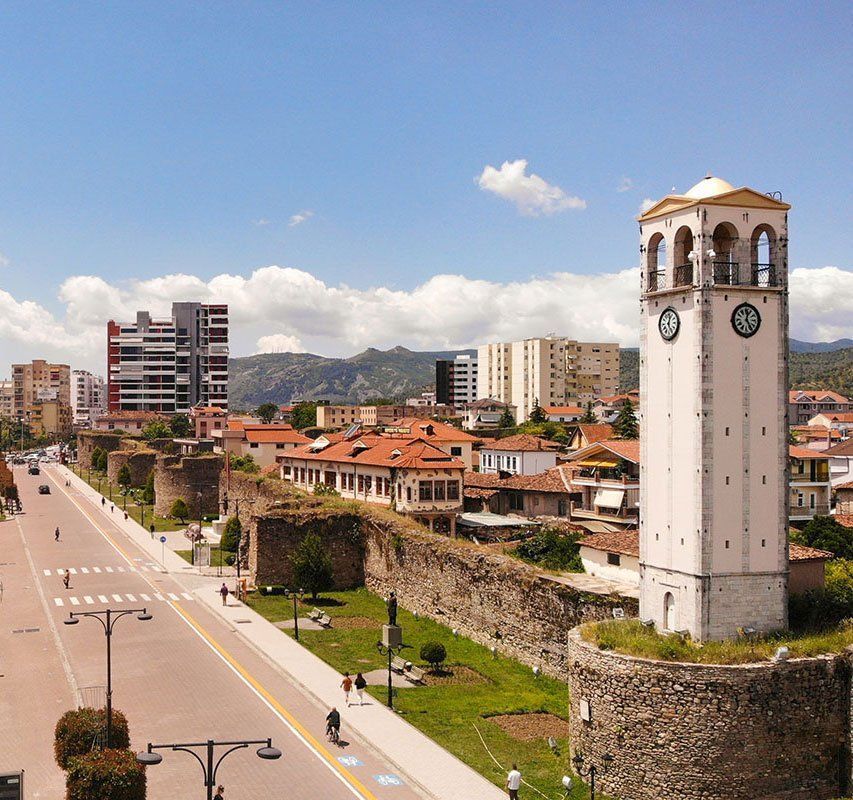 Clock tower overlooking city walls and a wide boulevard in Gjirokaster, Albania. Sunny day with blue sky and mountains in the background.