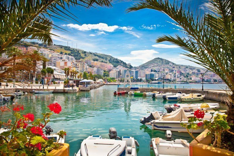 Boats docked in a turquoise harbor with a city on the hillside, palm trees and bright flowers in the foreground.