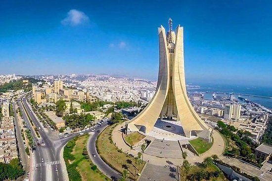 The Martyrs' Memorial in Algiers, Algeria. A large, beige monument with a central structure and three upward-curving arms, set against a city and sea backdrop.