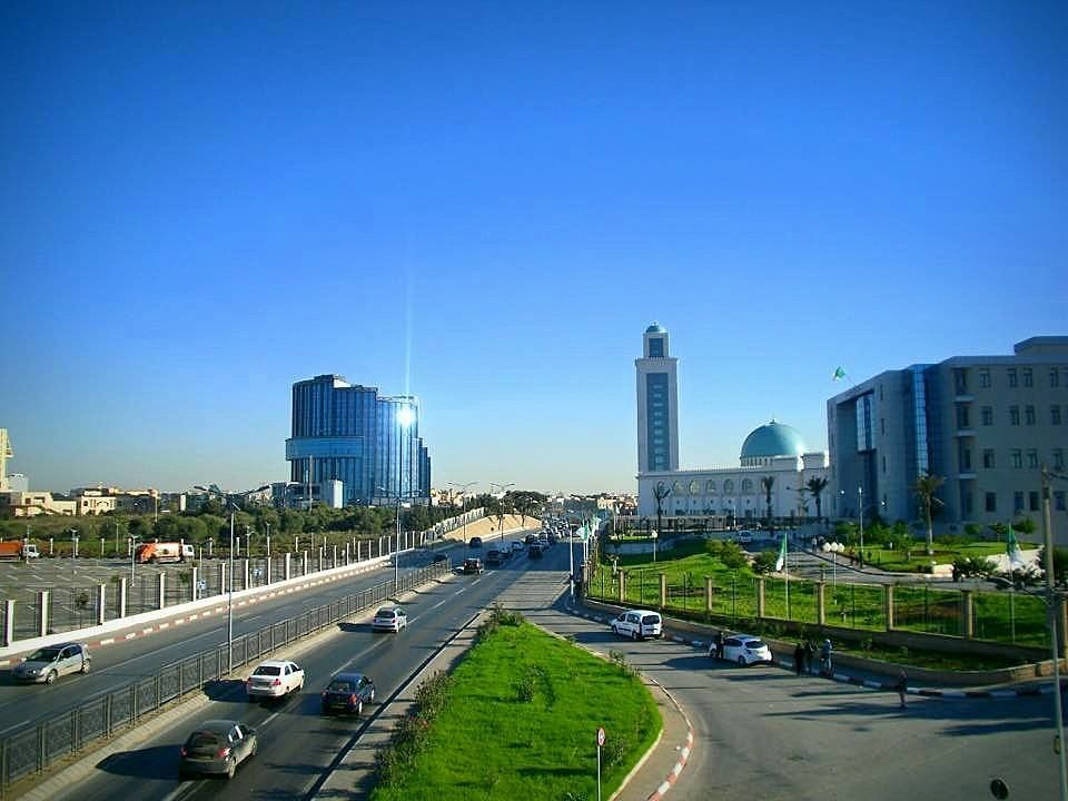 A sunny cityscape in Algeria featuring a highway, modern buildings, and a mosque with a tall minaret.
