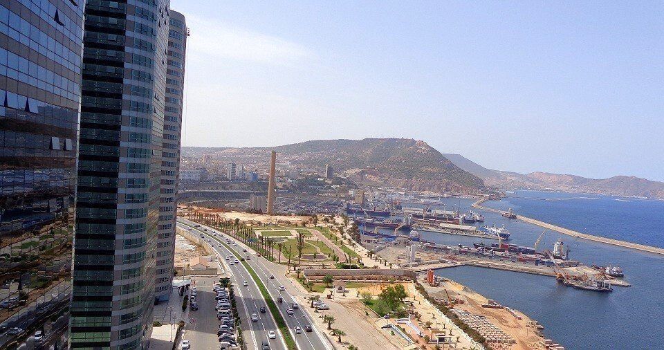 High-angle view of a coastal city with tall glass buildings, a highway, harbor, and a mountain in the background under a blue sky.