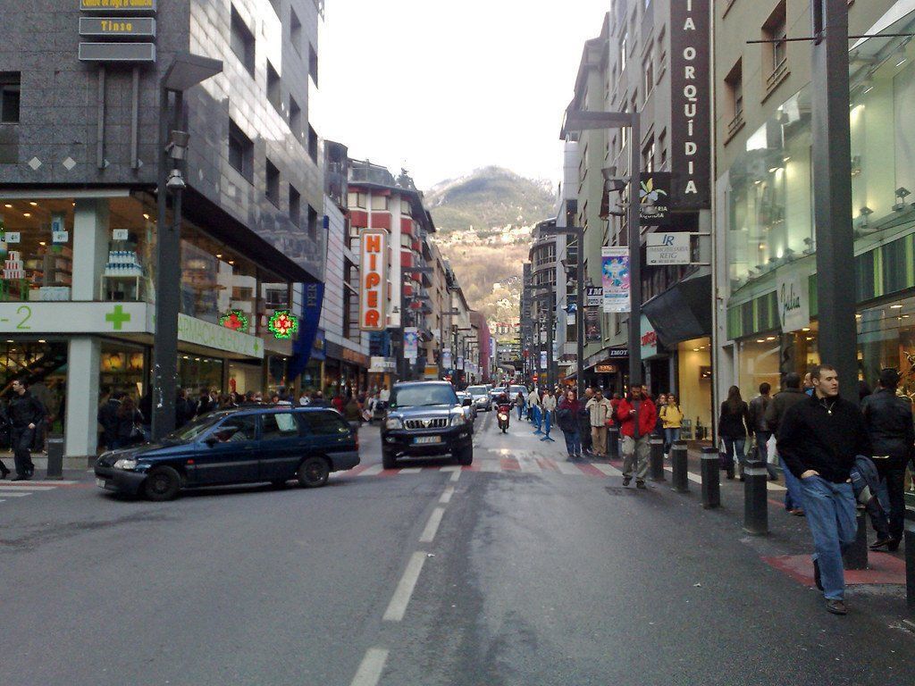Street scene in Andorra la Vella with shops, pedestrians, and cars. Buildings line the street with a mountain in the background.