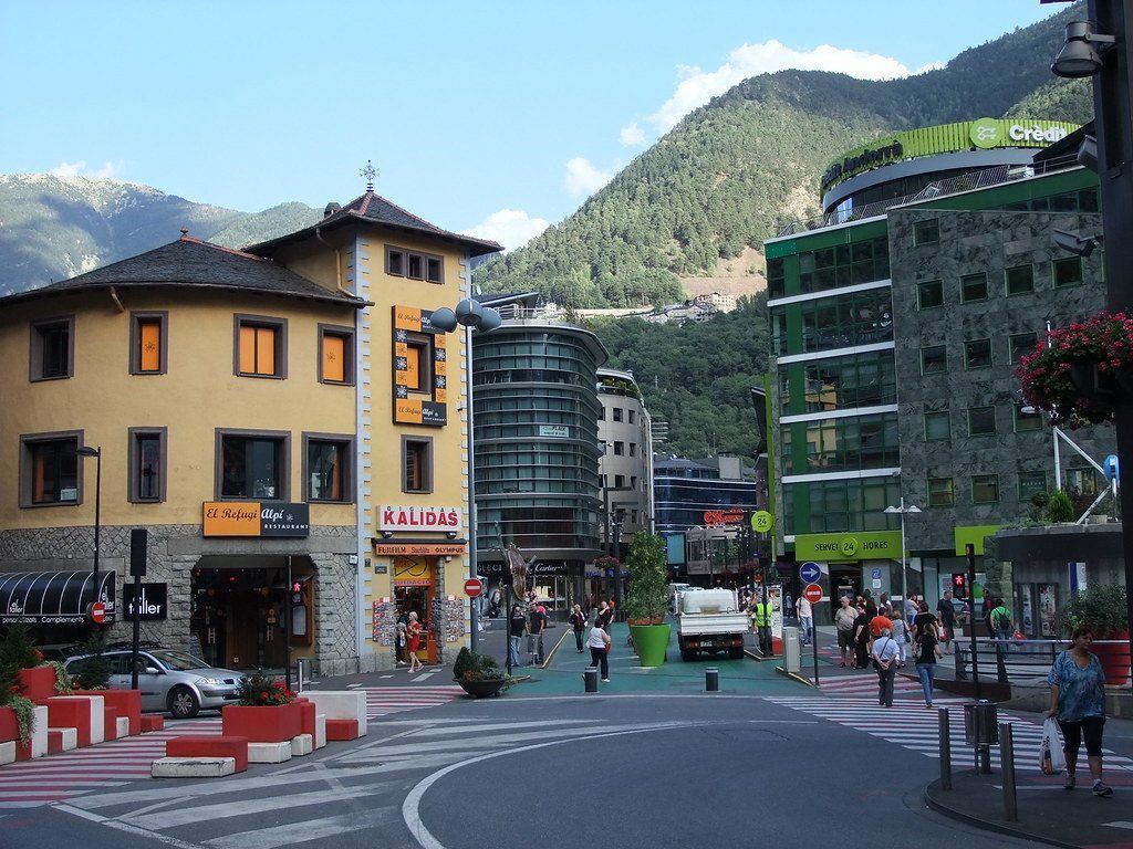 Street scene in Andorra la Vella, with buildings of various architectural styles, people walking, and mountains in the background.