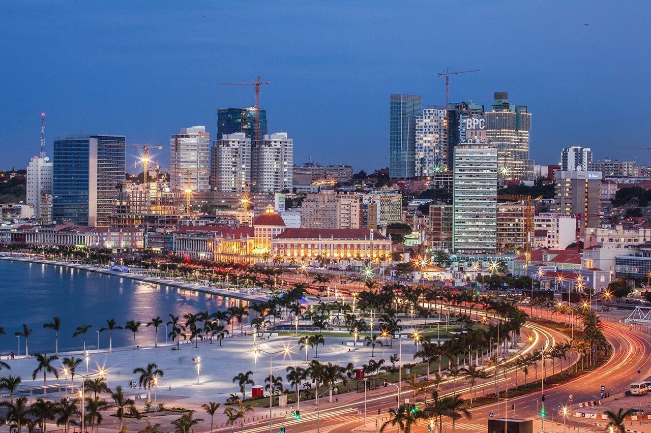 A nighttime cityscape of Luanda, Angola, with brightly lit buildings along a waterfront. Palm trees and illuminated roadways are in the foreground.