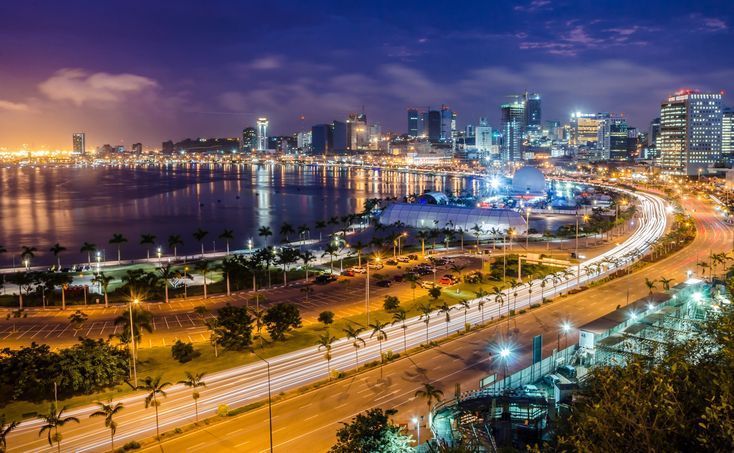 Nighttime cityscape of Luanda, Angola, with illuminated buildings along the waterfront and a highway with car lights.