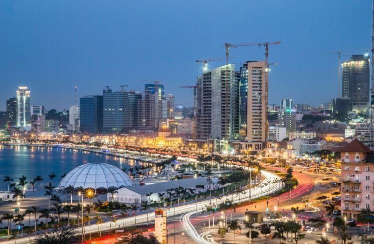 Nighttime view of Luanda, Angola, with a brightly lit city skyline along the waterfront, including high-rise buildings and a curved road with light trails.