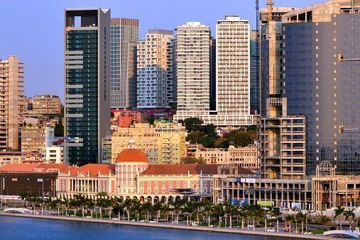 City skyline with modern skyscrapers and older buildings along a waterfront. The scene is bathed in warm sunlight.