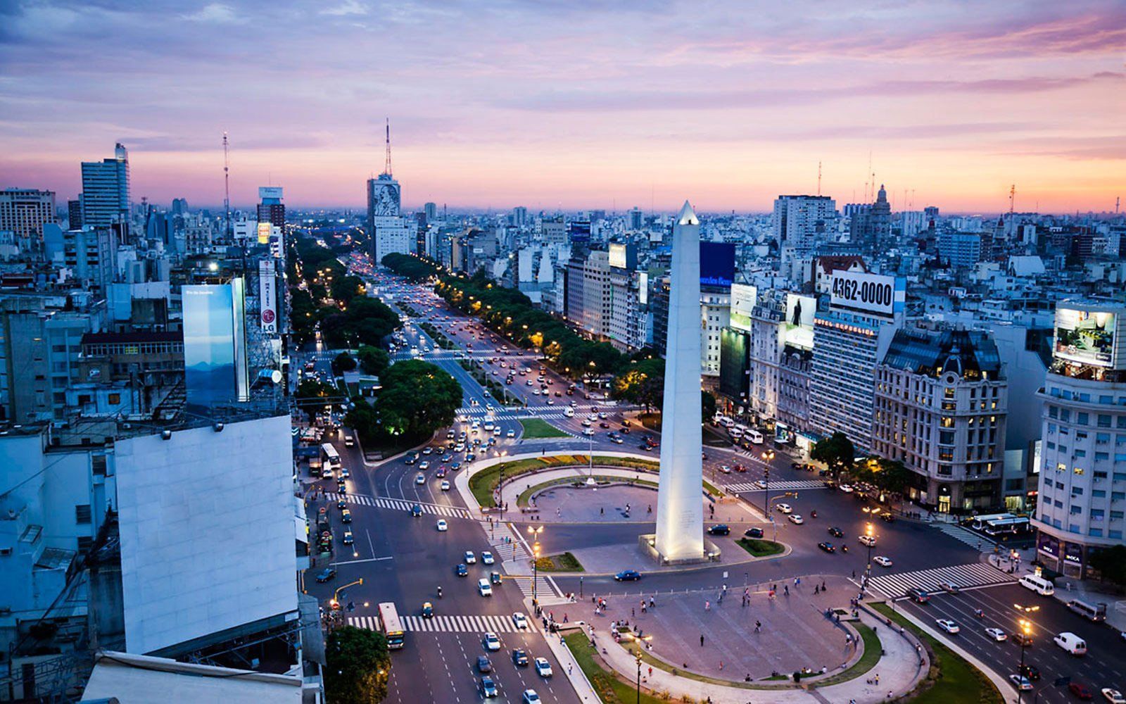 View of Buenos Aires, Argentina, at dusk with the Obelisco de Buenos Aires in the foreground. City lights and a colorful sky are visible.