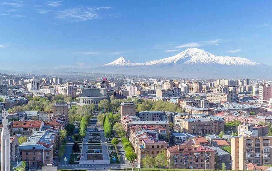 Cityscape of Yerevan, Armenia, with Mount Ararat in the background under a blue sky. Buildings and a tree-lined park are visible.