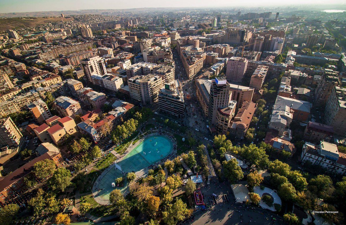 Aerial view of Yerevan, Armenia, with a central park featuring a blue water feature, surrounded by buildings and trees.