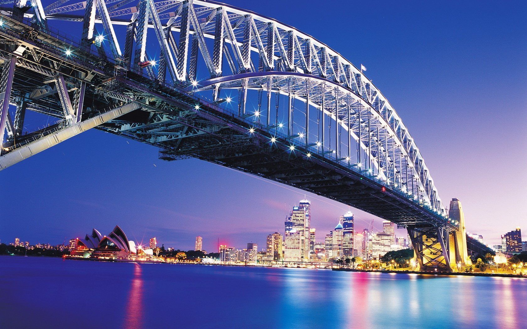 Sydney Harbour Bridge at night, with the city skyline in the background under a dark blue sky, illuminated by lights.