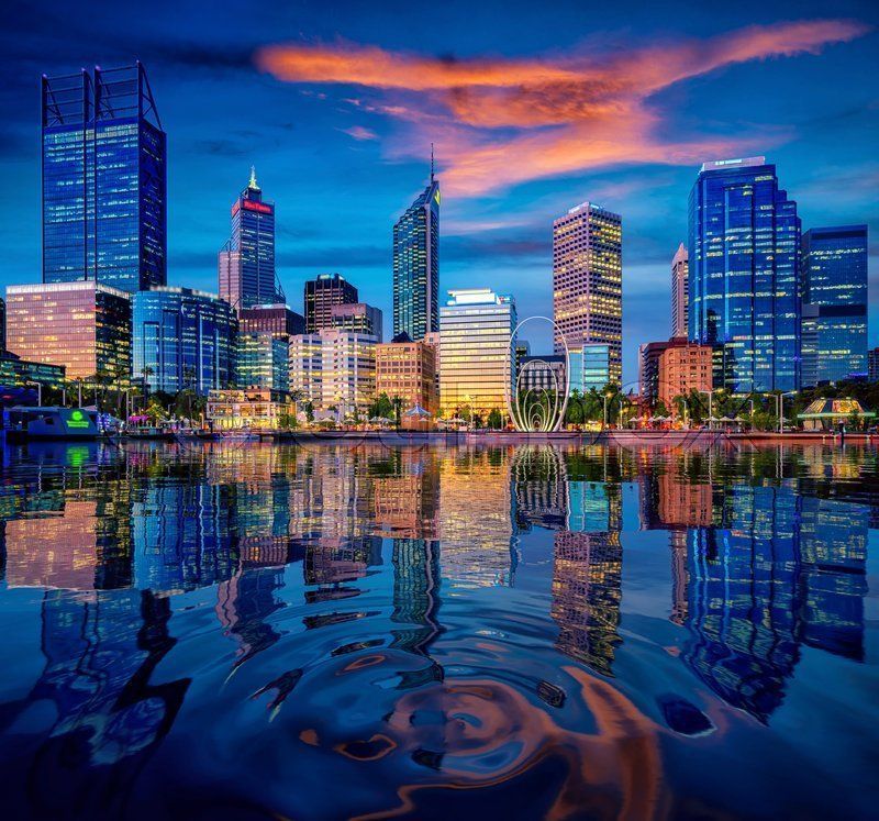 Perth, Australia skyline at dusk, reflected in calm water. Buildings lit with orange and blue lights, sunset sky.