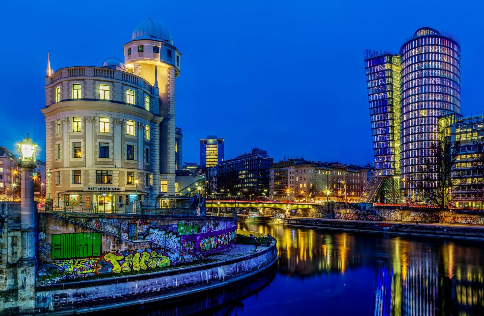 Nighttime cityscape with river. Historic tower and modern skyscraper glow against a dark blue sky. Graffiti on a riverbank wall.