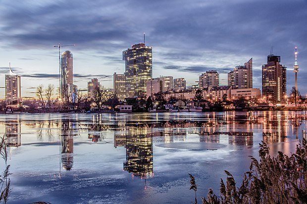 Vienna skyline at dusk reflected in calm water. Illuminated buildings and a distant tower.