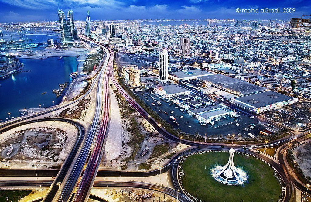 Aerial view of a brightly lit city at night with highways, skyscrapers, and a large roundabout with a monument.