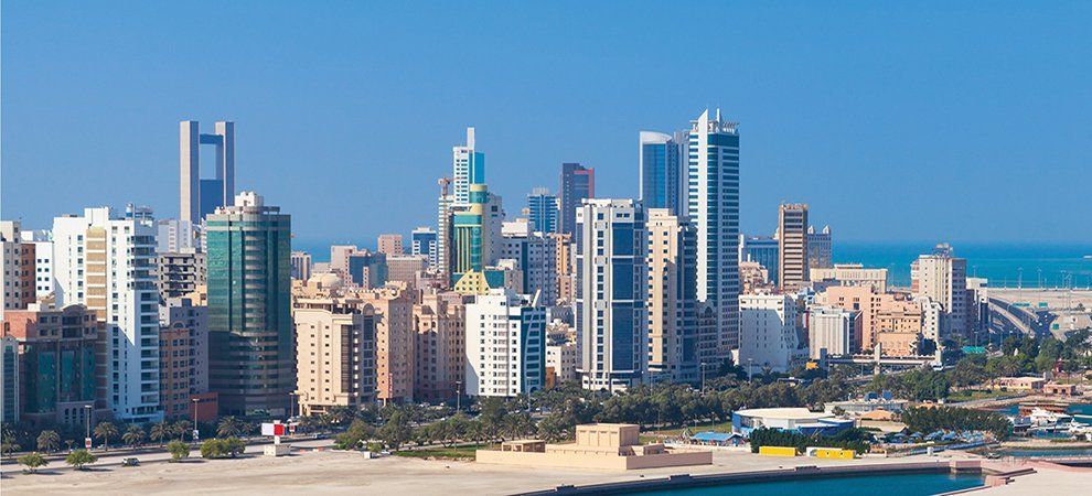 Skyline of a city with tall buildings, palm trees, and a body of water under a clear blue sky.