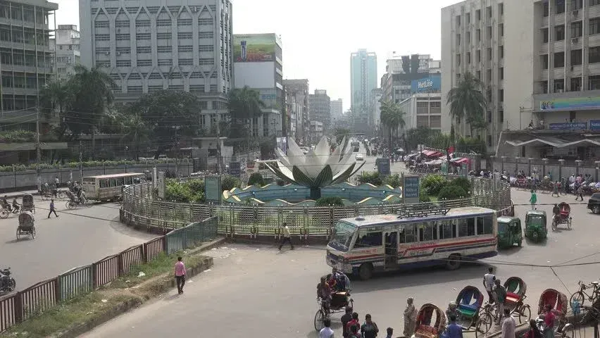 A busy city intersection in Dhaka, Bangladesh, with a large white lotus sculpture in the center, surrounded by traffic and pedestrians.