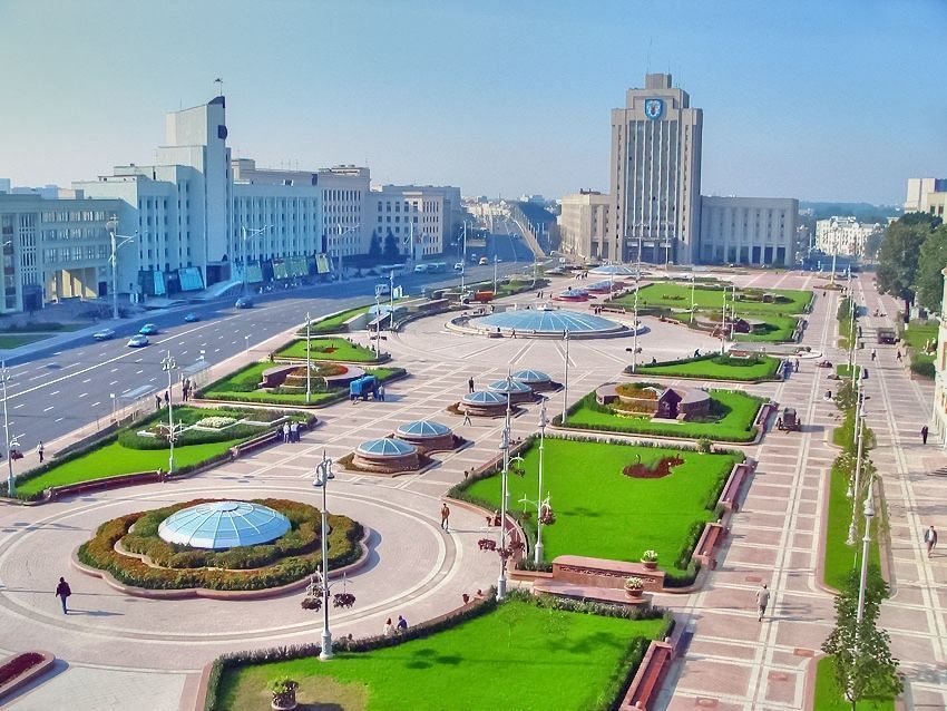 A large, manicured square in Minsk, Belarus, featuring green lawns, fountains, and grand white buildings under a blue sky.