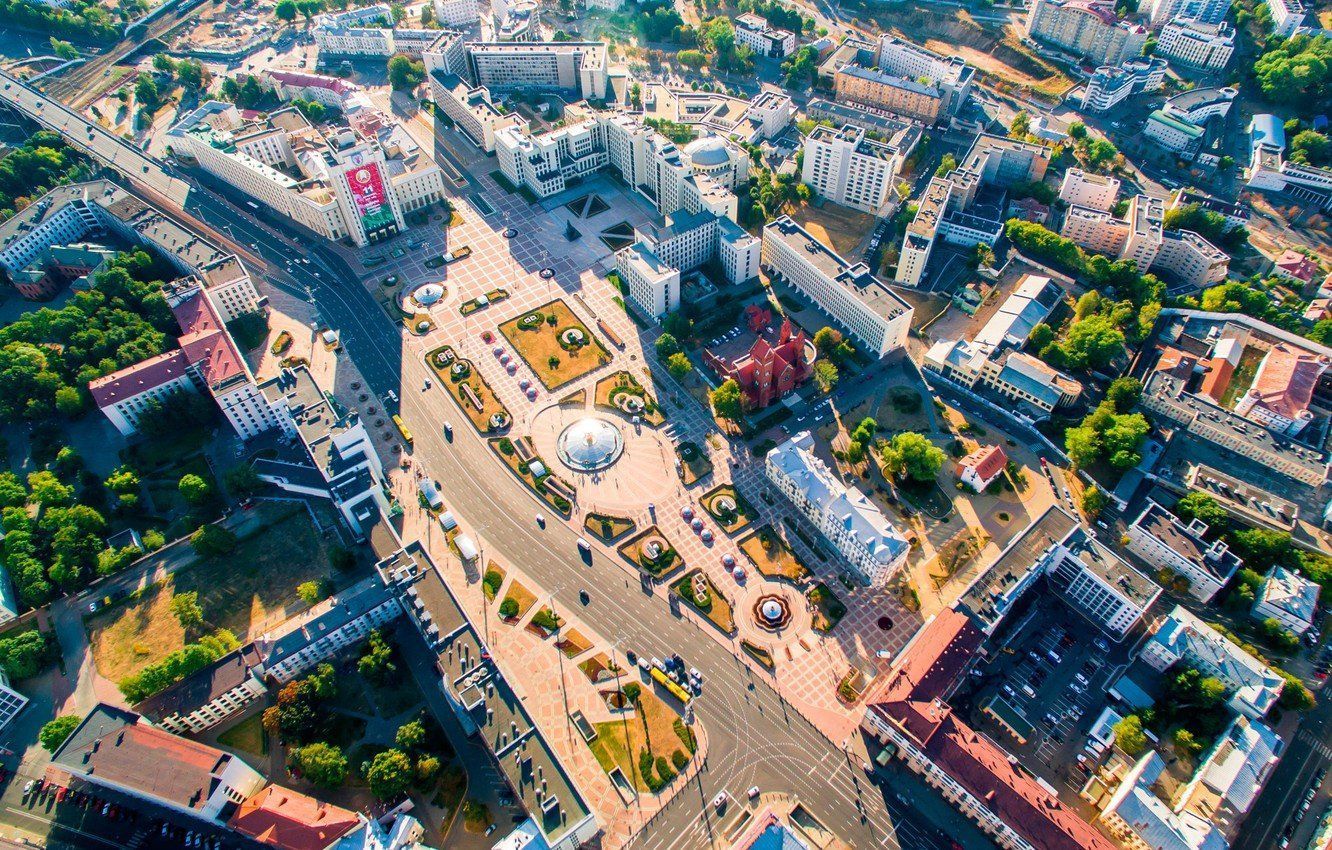 Aerial view of a city square with buildings, trees, and roads. A central plaza has a white structure surrounded by a circular walking path.