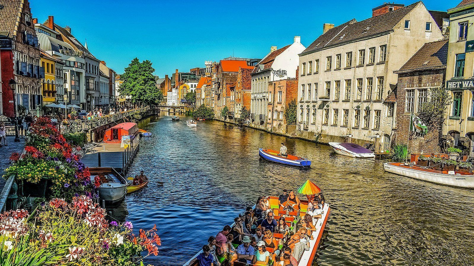 Canal with colorful boats, lined with buildings in a European city, with flowers on the banks.