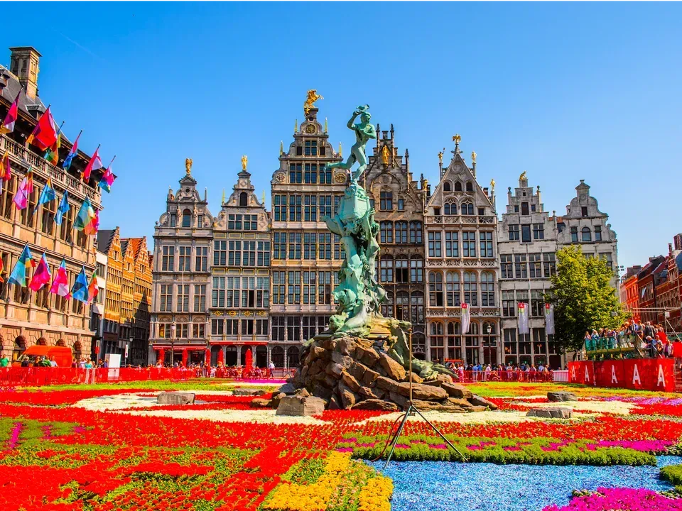 Grote Markt square in Antwerp, Belgium, with ornate guildhalls and a fountain sculpture, surrounded by a vibrant flower display under a blue sky.